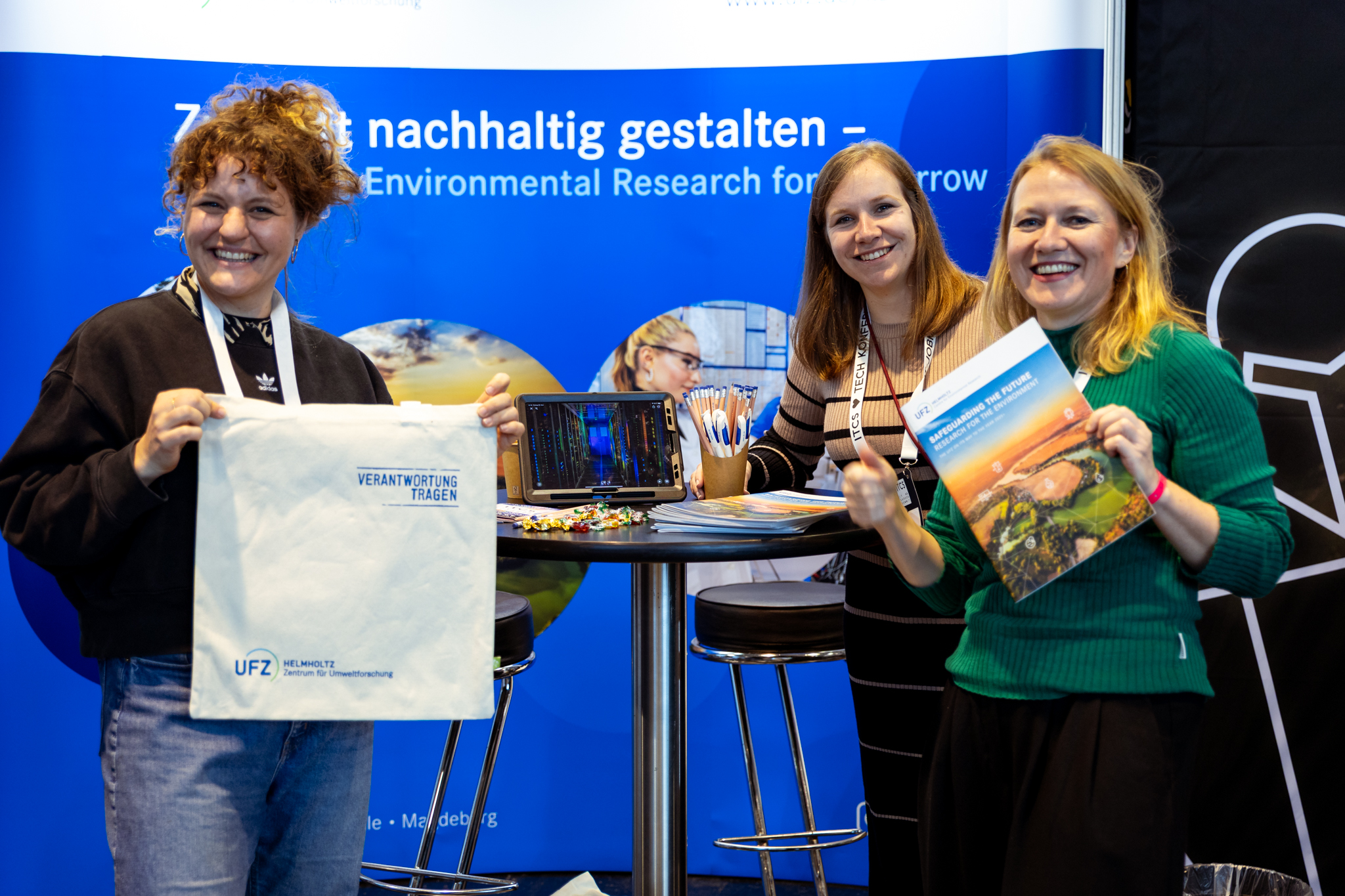 Visit to the ITCS trade fair in Frankfurt. Three UFZ employees are standing at the UFZ booth holding up merchandise.