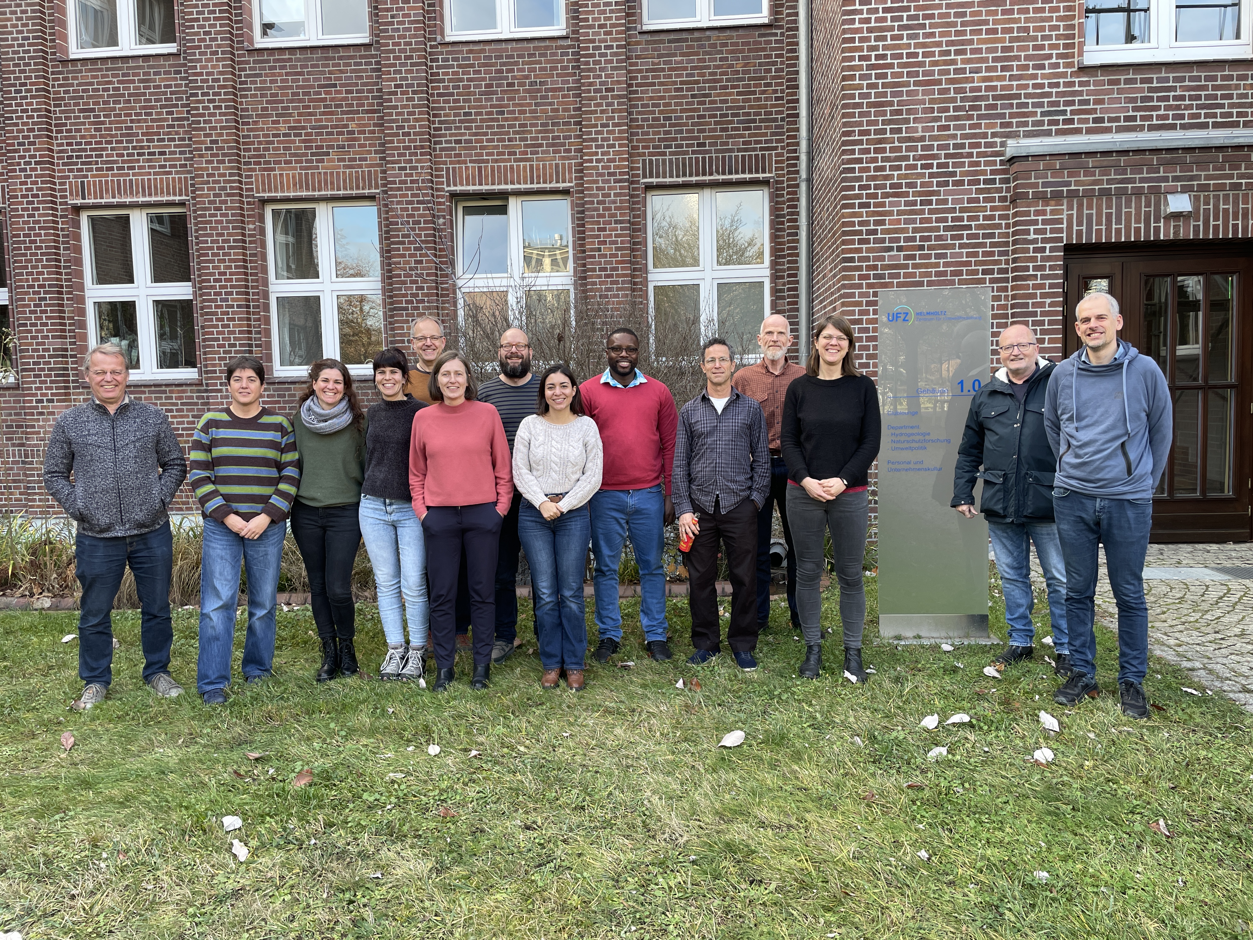 Ein Team aus 14 Wissenschaftler*innen steht auf einer Wiese vor einem Backsteingeb&auml;ude und l&auml;chelt in die Kamera. Zwischen den Personen steht ein hohes Hinweisschild aus Glas auf dem das UFZ-Logo und der Schriftzug Geb&auml;ude 1.0 zu sehen sind.