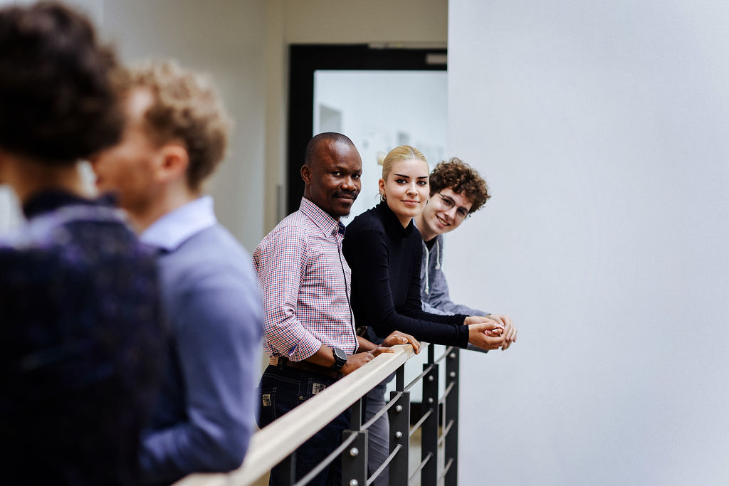 A small group of diverse people are standing together in a corridor, chatting and smiling at the camera. Photo: Anne Schwerin